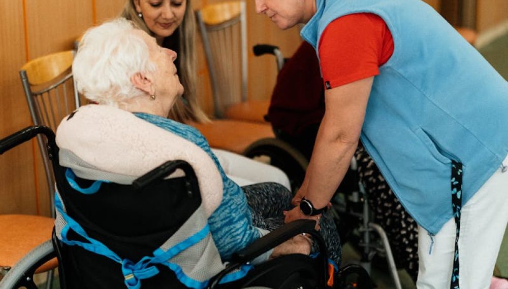 A caregiver talks with elderly women in a wheelchair at a retirement home.