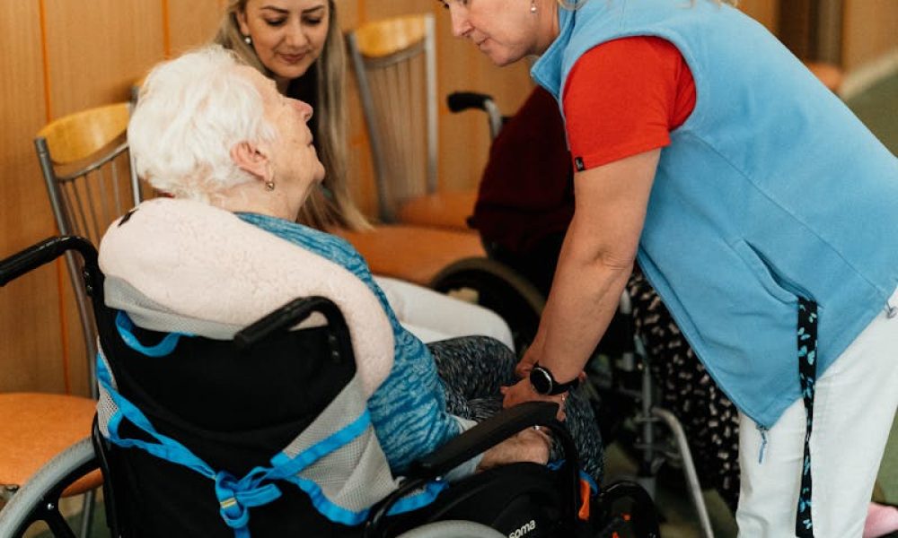 A caregiver talks with elderly women in a wheelchair at a retirement home.